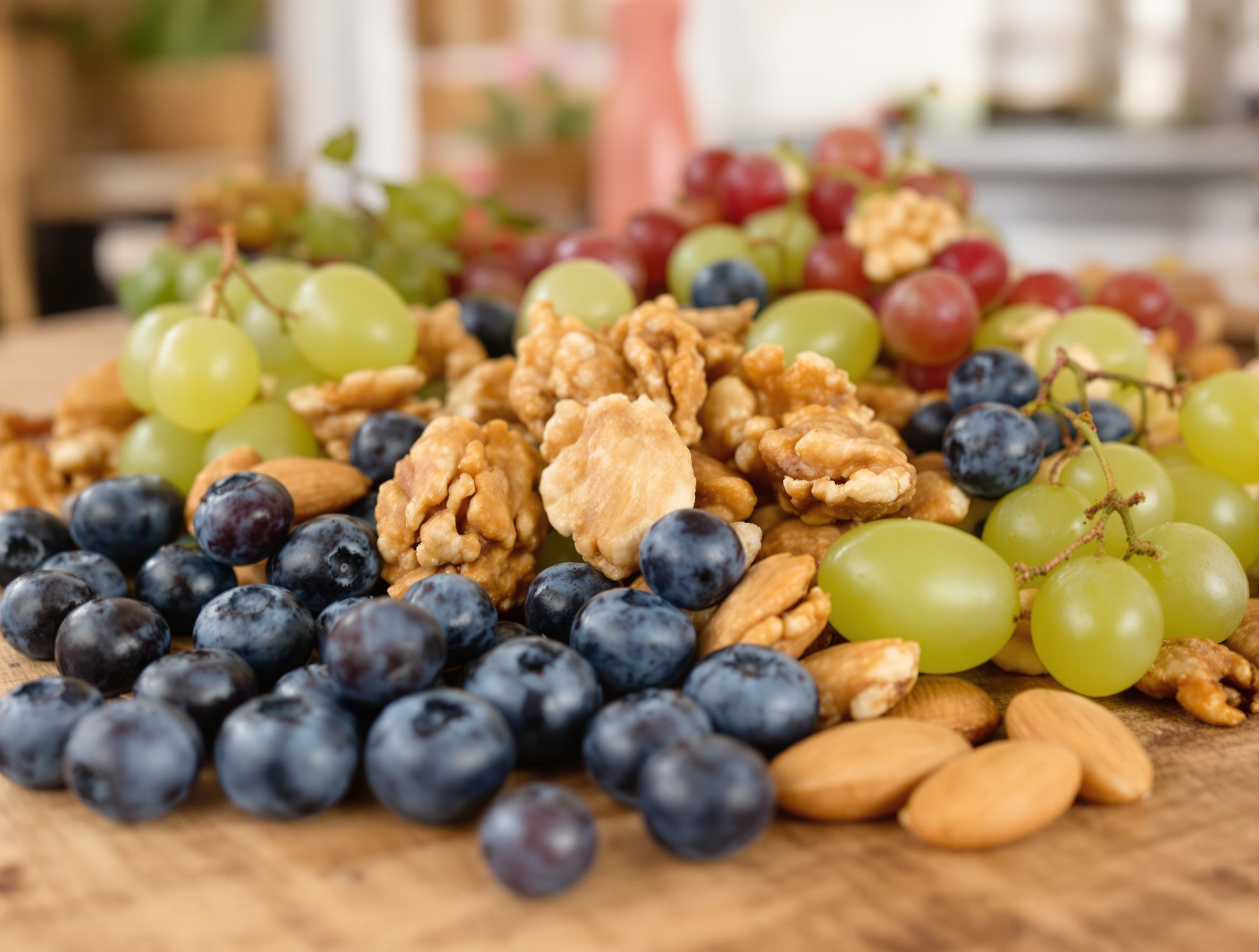 A close-up of fresh fruits and nuts, including blueberries, walnuts, almonds, and green grapes, arranged on a rustic wooden surface. The textures and colors are vibrant and detailed, with a softly blurred background.